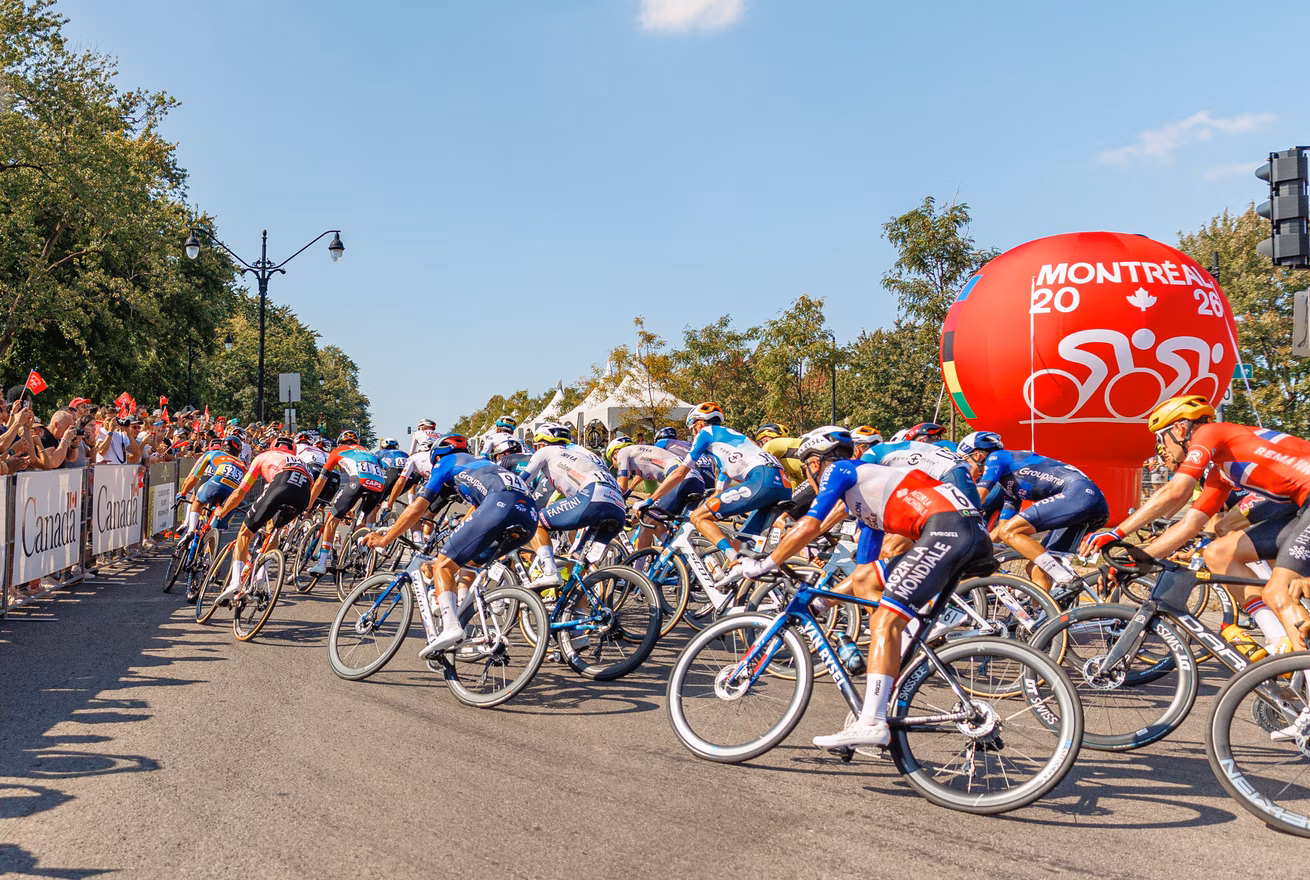 Des cyclistes professionnels tournent sur l'avenue du Parc à Montréal pendant les Grands Prix Cyclistes de Montréal 2024 devant un affichage promotionnel des Mondiaux 2026